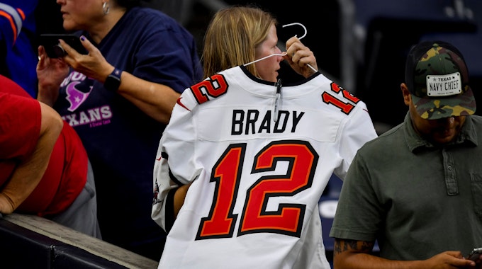 Ein Frau hält einen Kleiderbügel in den Händen. Daran hängt ein weißes Trikot von Tom Brady mit der Nummer 12.