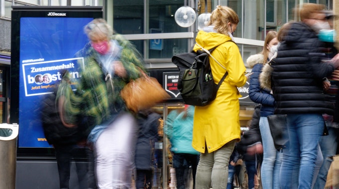 Einkaufstrubel zwischen den Jahren: Ein Bildschirm in einer Einkaufsstraße in der Kölner Innenstadt wirbt Ende Dezember für die Boosterimpfung. Das RKI schlüsselt bei einem kleinen Teil der Neuinfektionen auf, an welchen Orten sie stattfinden.