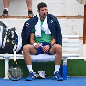 Novak Djokovic sitzt mit Jacke und Handtuch auf einer Bank, neben ihm sein Tennisschläger.