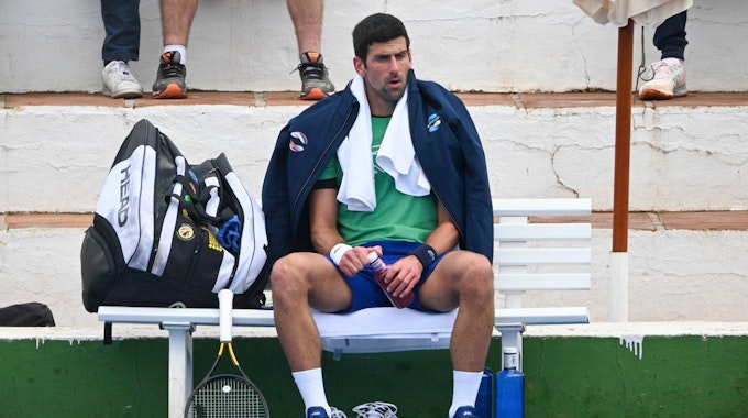 Novak Djokovic sitzt mit Jacke und Handtuch auf einer Bank, neben ihm sein Tennisschläger.