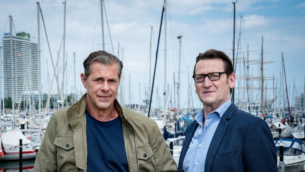 Sven Martinek (l.) und Ingo Naujoks stehen vor einem Hafen.