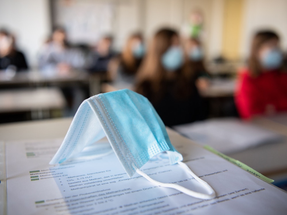 Eine Maske liegt im Unterricht auf Unterlagen, während im Hintergrund Schülerinnen und Schüler mit Mund- und Nasenschutz zu sehen sind.