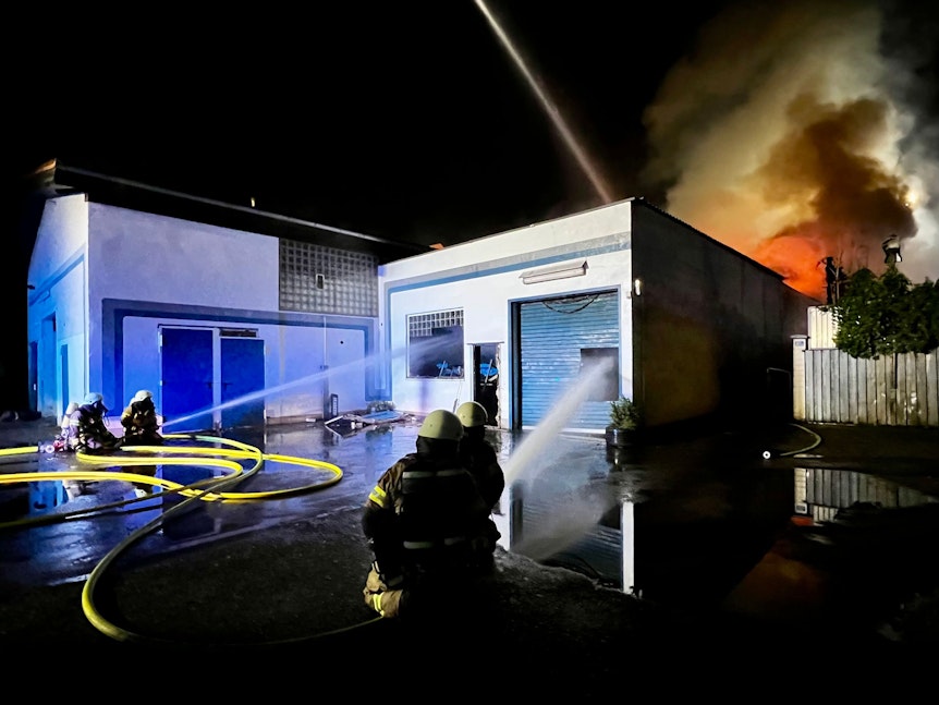 Feuerwehrleute löschen mit Wasser in eine Lagerhalle, im Hintergrund die Flammen.