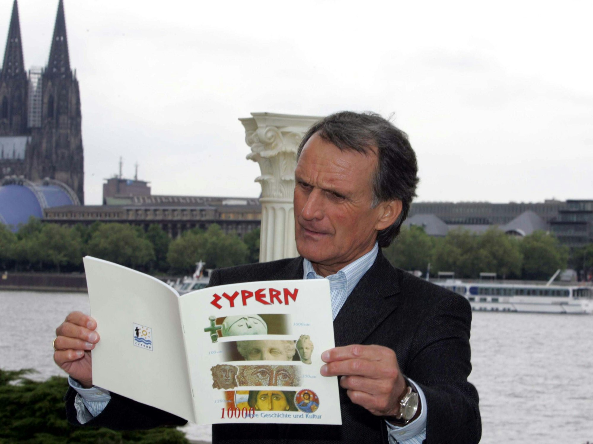 Wolfang Overatsch hält eine Broschüre über Zypern in den Händen. Im Hintergrund ist der Rhein samt Dom zu sehen.