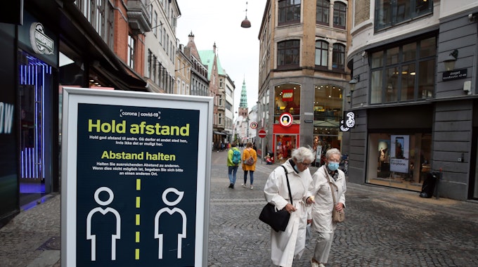 „Abstand halten“ steht auf einem Schild in einer Straße in Kopenhagen, Passanten laufen daran vorbei.