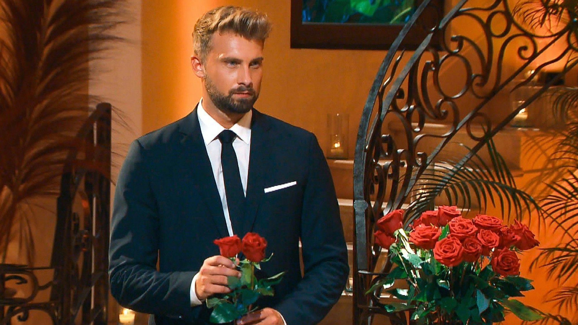 „Bachelor“ Dominik Stuckmann in einem schwarzen Anzug auf einem Foto aus der Show am 26. Januar 2022. In der Hand hält er zwei Rosen.