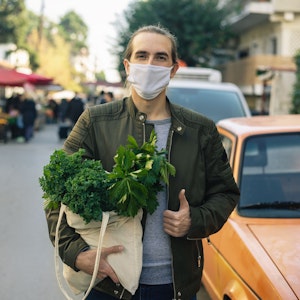 Millennial man with cloth face mask standing outdoor with reusable bag with vegetables inside and street local farmers market behind him