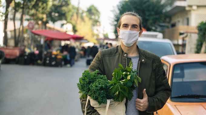 Millennial man with cloth face mask standing outdoor with reusable bag with vegetables inside and street local farmers market behind him