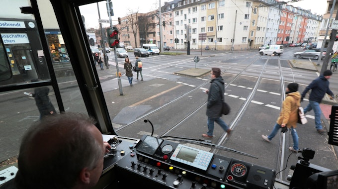 Blick aus dem Führerstand einer KVB-Bahn auf die Straße, wo Menschen die Gleise überqueren.