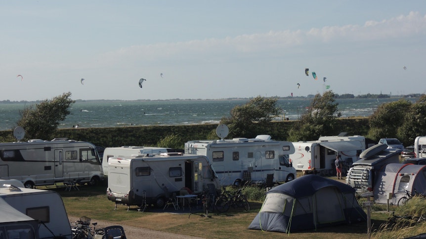 Wohnmobile stehen auf einem Campingplatz. Im Hintergrund sieht man das Meer.