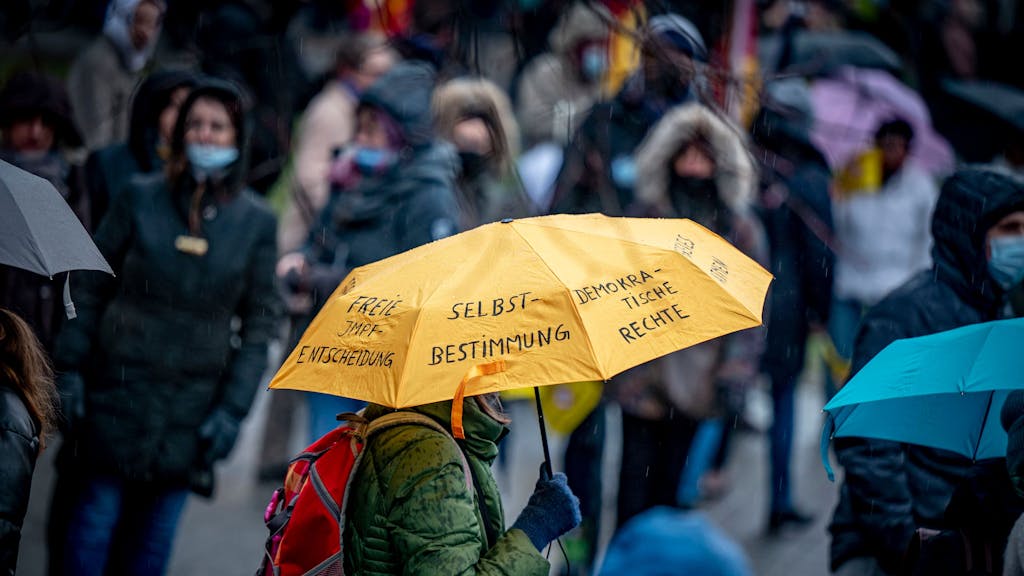 Gegner der Corona-Maßnahmen nehmen an einer Demonstration teil, eine Person hält einen Regenschirm, auf dem u.&nbsp;a. „Selbstbestimmung“ und „Demokratische Rechte“ steht.