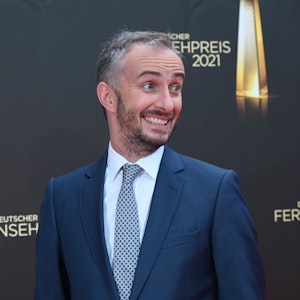 Jan Böhmermann kommt zur Verleihung des Deutschen Fernsehpreises 2021 im Tanzbrunnen. Der Satiriker steht vor einer Wand mit dem Logo der Veranstaltung und grinst schief an der Kamera vorbei.