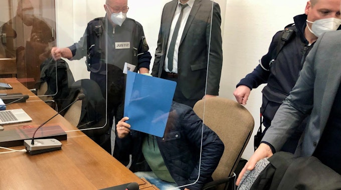 Ein Mann sitzt im Gericht auf der Anklagebank und hält sich eine Mappe vor das Gesicht.