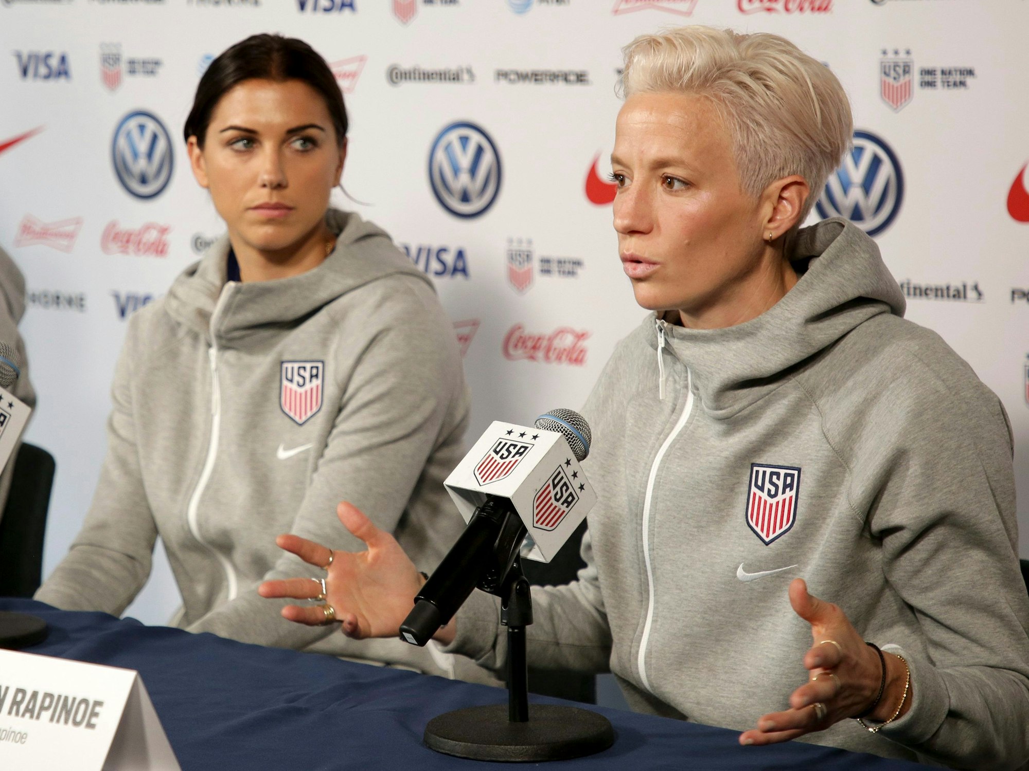Frauen-Nationalmannschaft der USA bei einer Pressekonferenz. Megan Rapinoe spricht ins Mikrofon. Team-Kollegin Alex Morgan schaut sie an.
