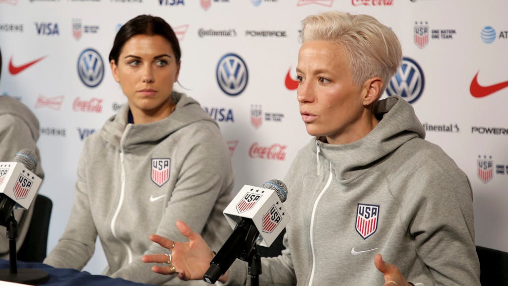Frauen-Nationalmannschaft der USA bei einer Pressekonferenz. Megan Rapinoe spricht ins Mikrofon. Team-Kollegin Alex Morgan schaut sie an.