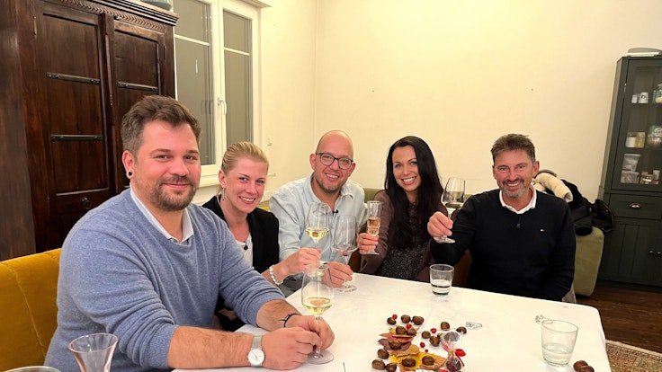 Die Das Perfekte Dinner-Kandidaten Marcus, Simone, Gastgeber Andreas, Sabrina und Jürgen sitzen an Andreas Tisch und lächeln.