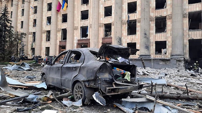 Ein zerstörtes Auto steht vor dem beschädigten Rathausgebäude von Charkiw