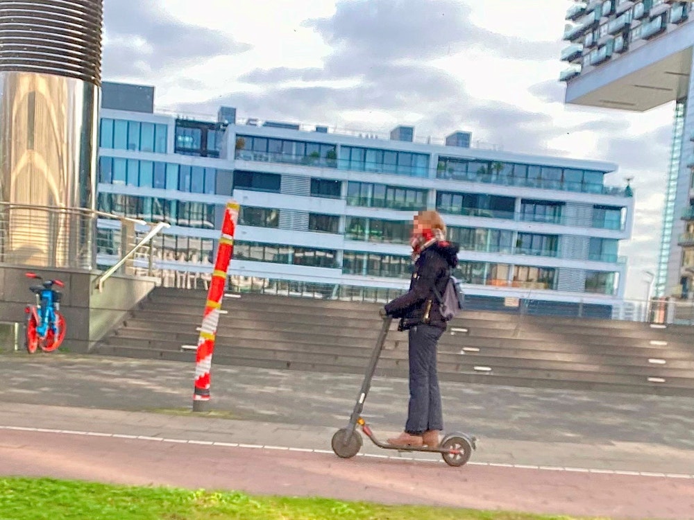 Ein Mast mitten auf dem Gehweg am Rheinauhafen in Köln, fotografiert vor wenigen Tagen. Eine Person fährt mit einem Roller daran vorbei.