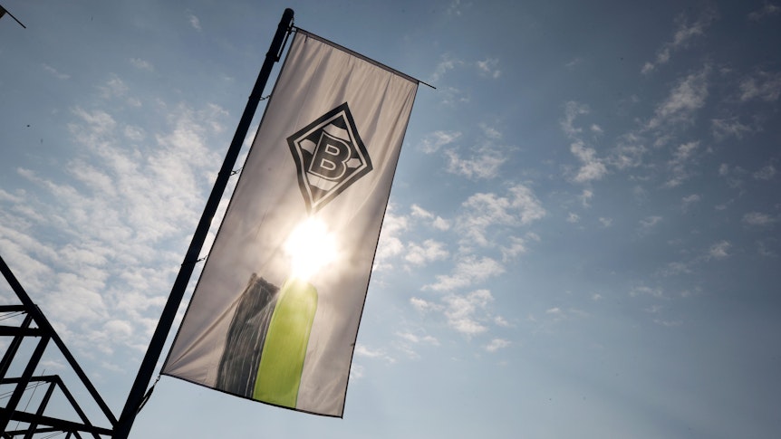 Eine Fahne mit dem Logo von Fußball-Bundesligist Borussia Mönchengladbach weht im Borussia-Park. Im Hintergrund scheint die Sonne. Dieses Foto stammt vom 7. April 2020.