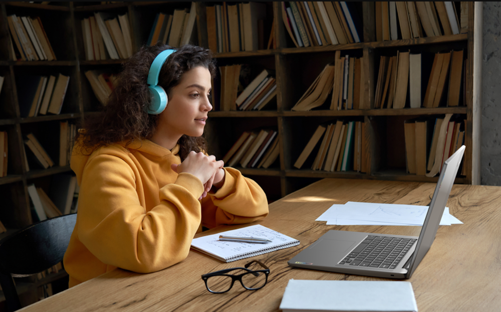 Eine Frau im gelben Pullover und Kopfhörern am Ohr sitzt am Tisch vor Ihrem Laptop.