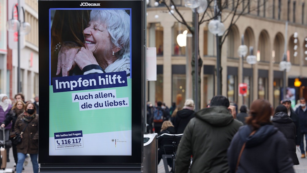 Menschen gehen an einer elektronischen Werbetafel mit der Aufschrift „Impfen hilft. Auch allen, die du liebst“ in der Innenstadt vorbei.
