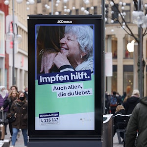 Menschen gehen an einer elektronischen Werbetafel mit der Aufschrift „Impfen hilft. Auch allen, die du liebst“ in der Innenstadt vorbei.