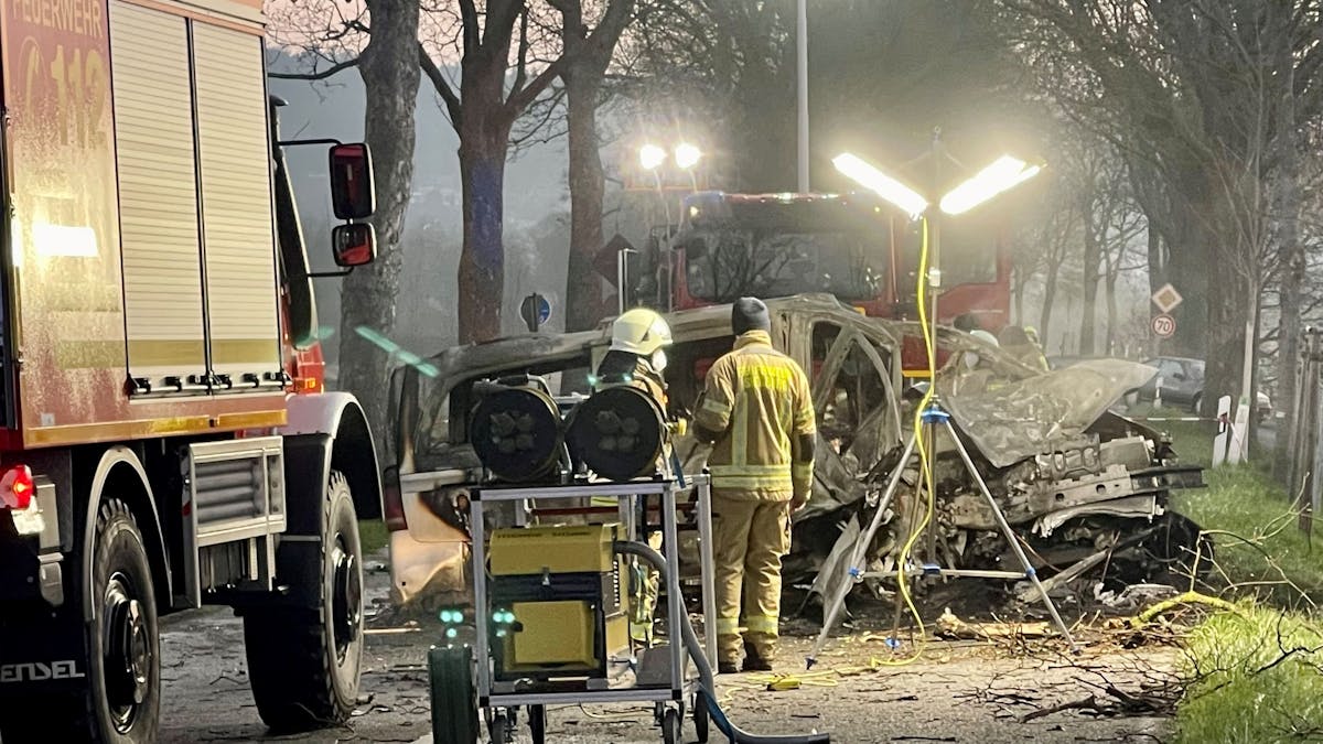 Zwei Feuerwehrmänner stehen vor dem ausgebrannten Wrack eines Transporters, in dem zwei junge Männer (beide 17) aus Siegburg gestorben sind.