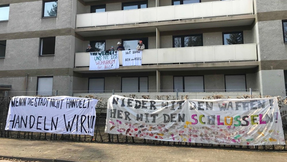 Vier Männer haben ein Gebäude besetzt, sie befinden sich auf einem der Balkone. Am Balkon und am Zaun hängen Banner mit der Aufschrift „Nein zum Krieg“ und „Nieder mit den Waffen! Her mit den Schlüsseln!“