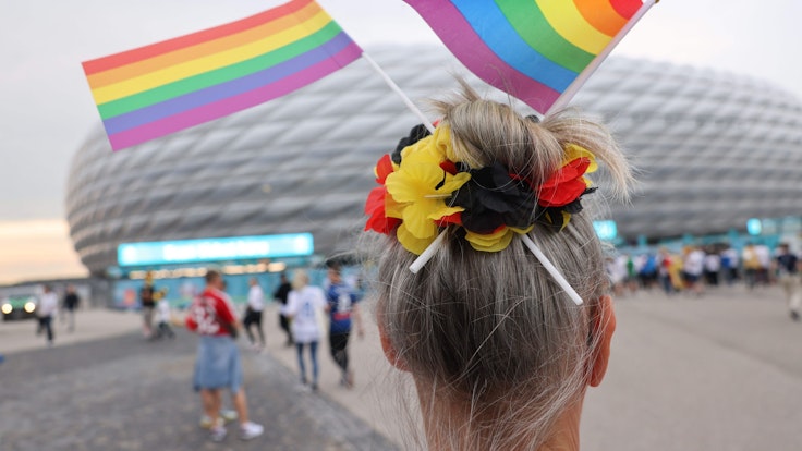 Eine Fußball-Anhängerin mit zwei kleinen Regenbogenfahnen im Haar steht vor der Münchner Allianz-Arena.