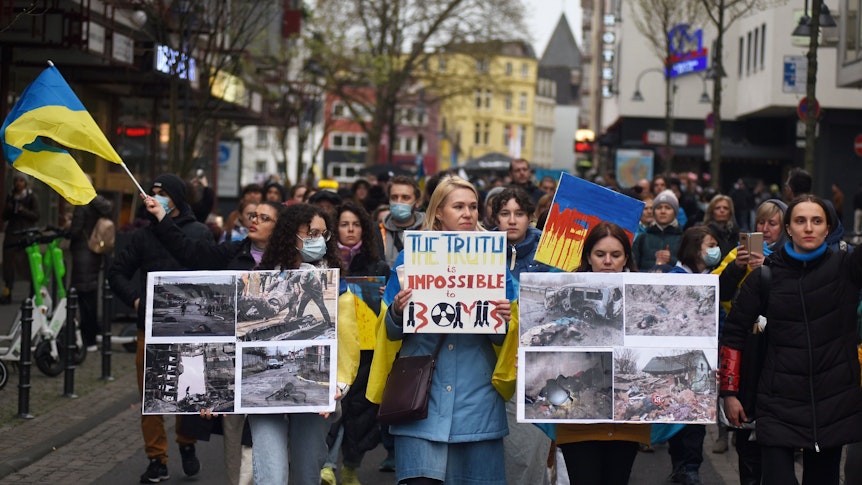 Teilnehmer einer Kundgebung laufen durch eine Straße, sie halten Plakate mit Fotos unter anderem von zerstörten Häusern in der Ukraine in Händen.