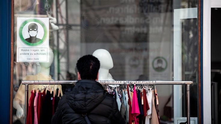 Eine Frau schaut sich Kleidung an einer Stange vor einem Geschäft an, auf einem Schild im Fenster steht "Maske tragen, um andere zu schützen".