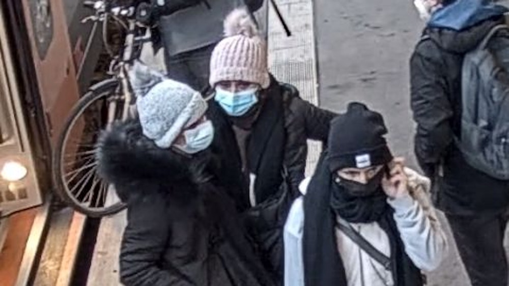 Drei junge Frauen stehen auf einem Bahnsteig zusammen. Alle drei tragen Schutzmasken und Wintermützen, eine von ihnen telefoniert.