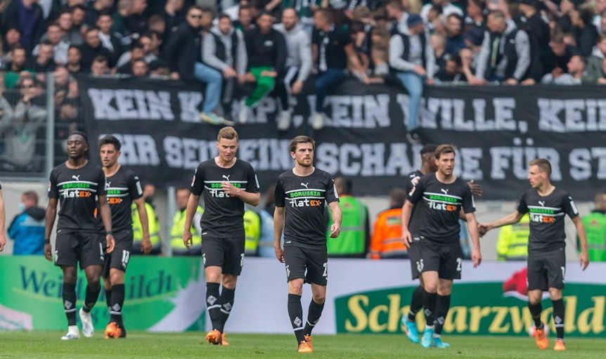 Die Spieler von Borussia Mönchengladbach schreiten nach dem 3:3 in Freiburg (23. April 2022) vom Rasen, im Hintergrund sind die Fohlen-Fans zu sehen. Am Zaun hängt ein Banner.