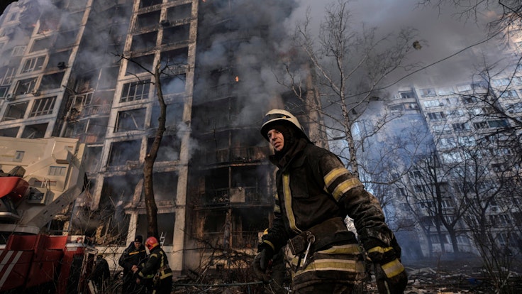 15.03.2022, Ukraine, Kyjiw: Einsatzkräfte der Feuerwehr stehen vor einem brennenden Wohngebäude, das durch Bombenbeschuss des russischen Militärs zerstört wurde.