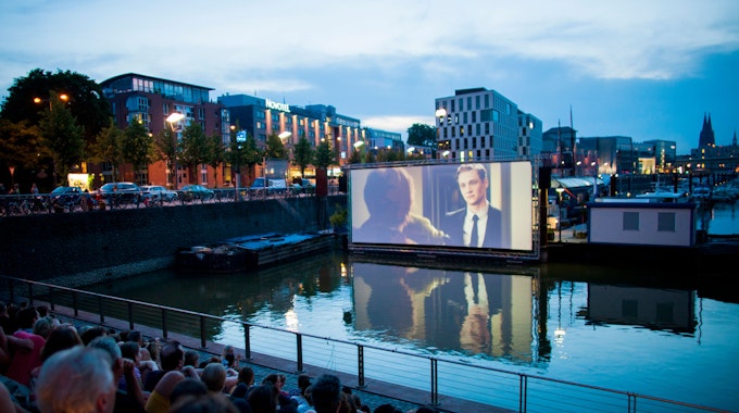 Menschen schauen sich im Open Air Kino am Rheinauhafen in Köln vor der Domkulisse einen Film an.