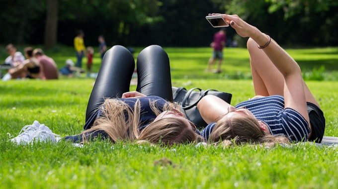Zwei jungen Frauen liegen am 12. Mai 2018 auf einer Wiese im Park, eine von den Frauen hält ein Smartphone in der Hand.