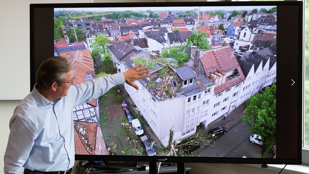 Der Bürgermeister von Paderborn, Michael Dreier, spricht bei einer Pressekonferenz am 21. Mai vor einem Display mit einem Schadensbild aus der Innenstadt, das zeigt, wie Bäume auf ein Hausdach geweht worden sind.