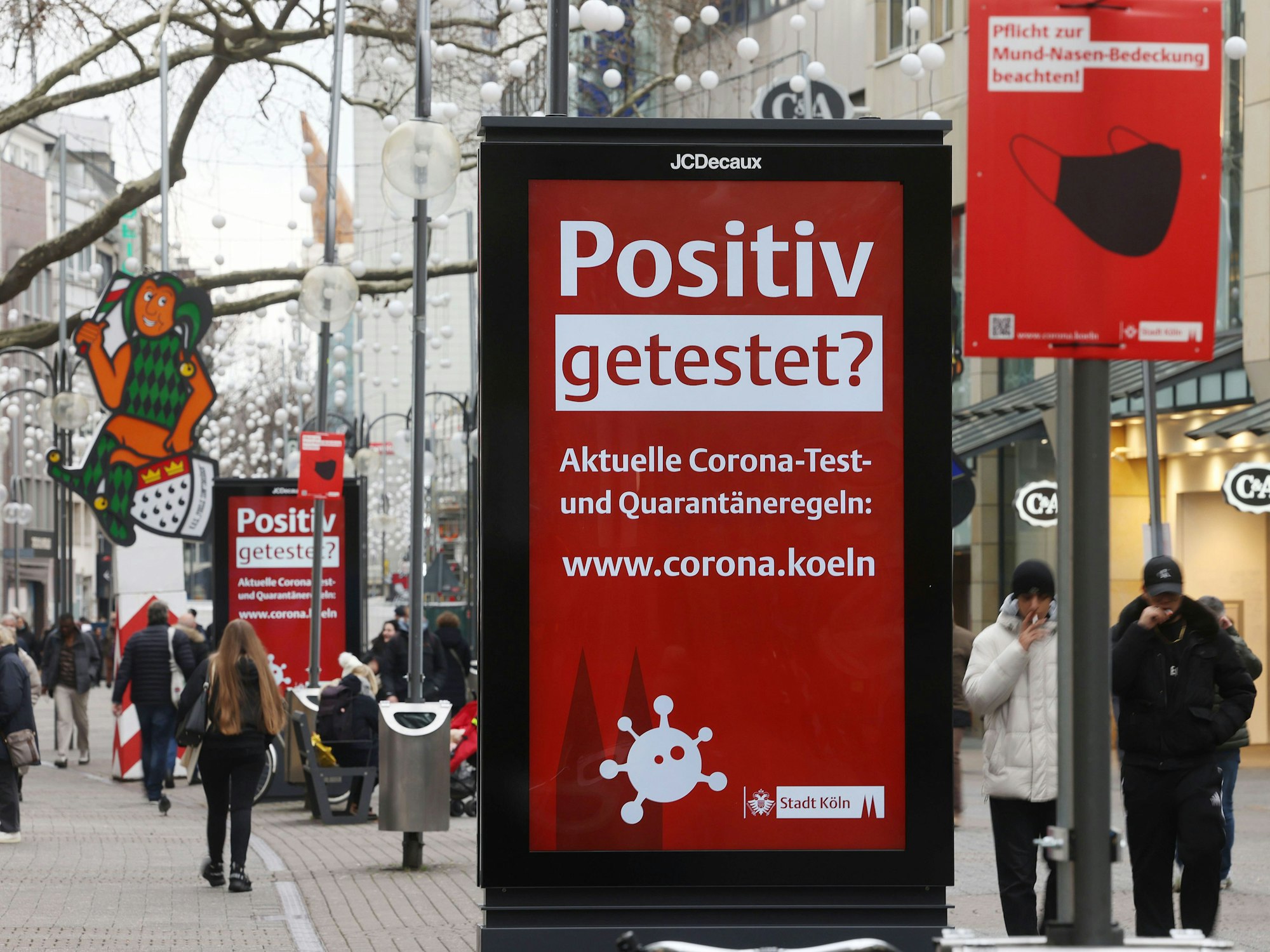 Menschen gehen an einer elektronischen Werbetafel mit der Aufschrift "Positiv getestet?" in der Kölner Innenstadt vorbei.