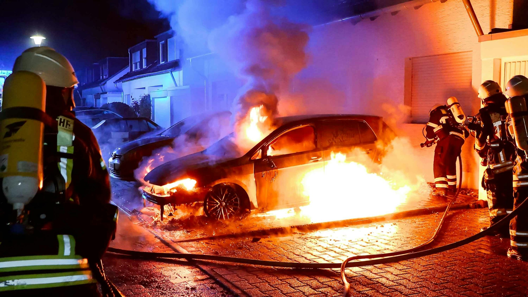Feuer in Köln: Ein Auto steht in Flammen, mehrere Feuerwehrmänner sind im Einsatz.