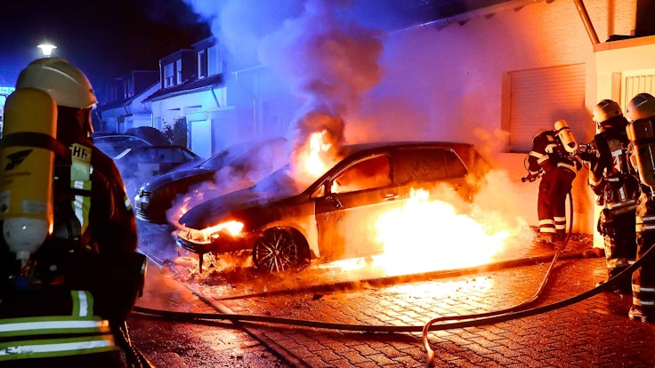 Ein Auto, das vor einem Haus steht, brennt lichterloh. Feuerwehrmänner löschen.