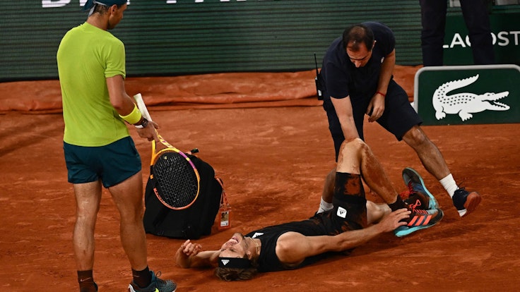 Alexander Zverev liegt schreiend am Boden, während ihn ein Arzt behandelt. Gegenspieler Rafael Nadal leidet daneben mit.