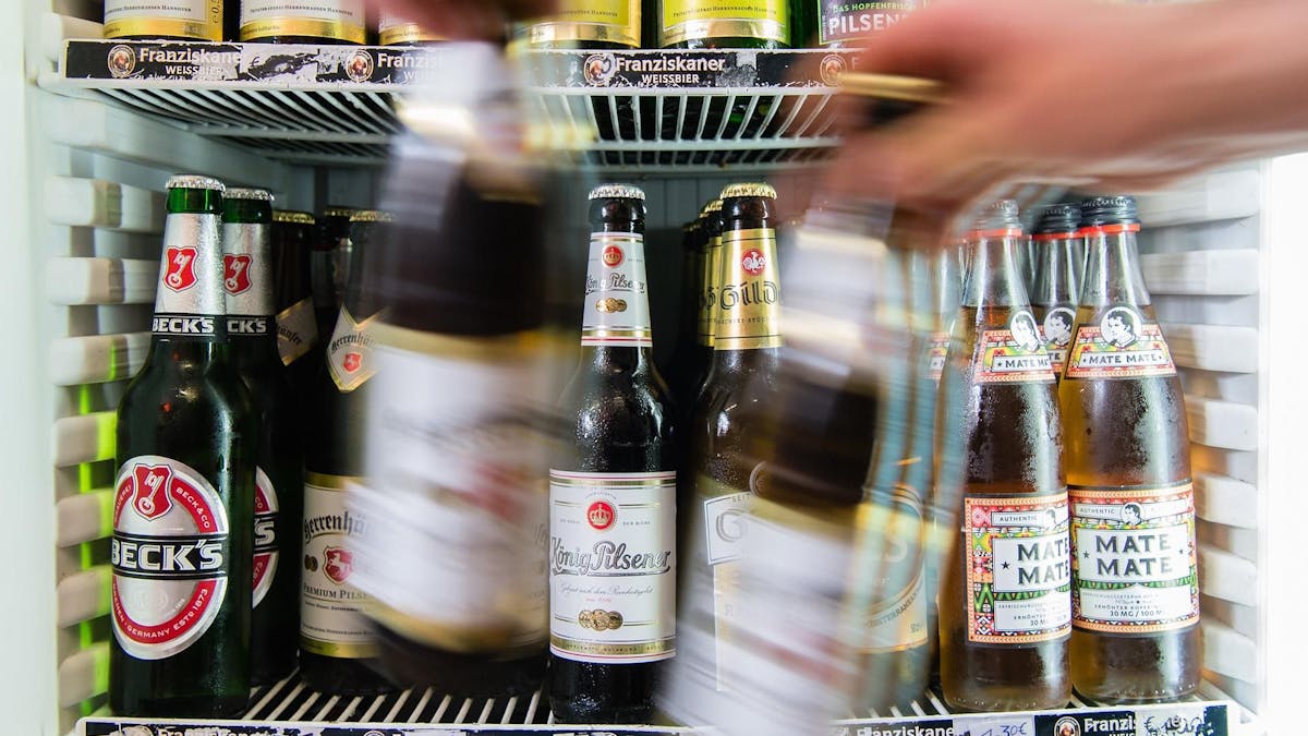 Zwei Personen nehmen sich ein Bier aus einem Kiosk. Im Hintergrund sind mehrere Bierflaschen.