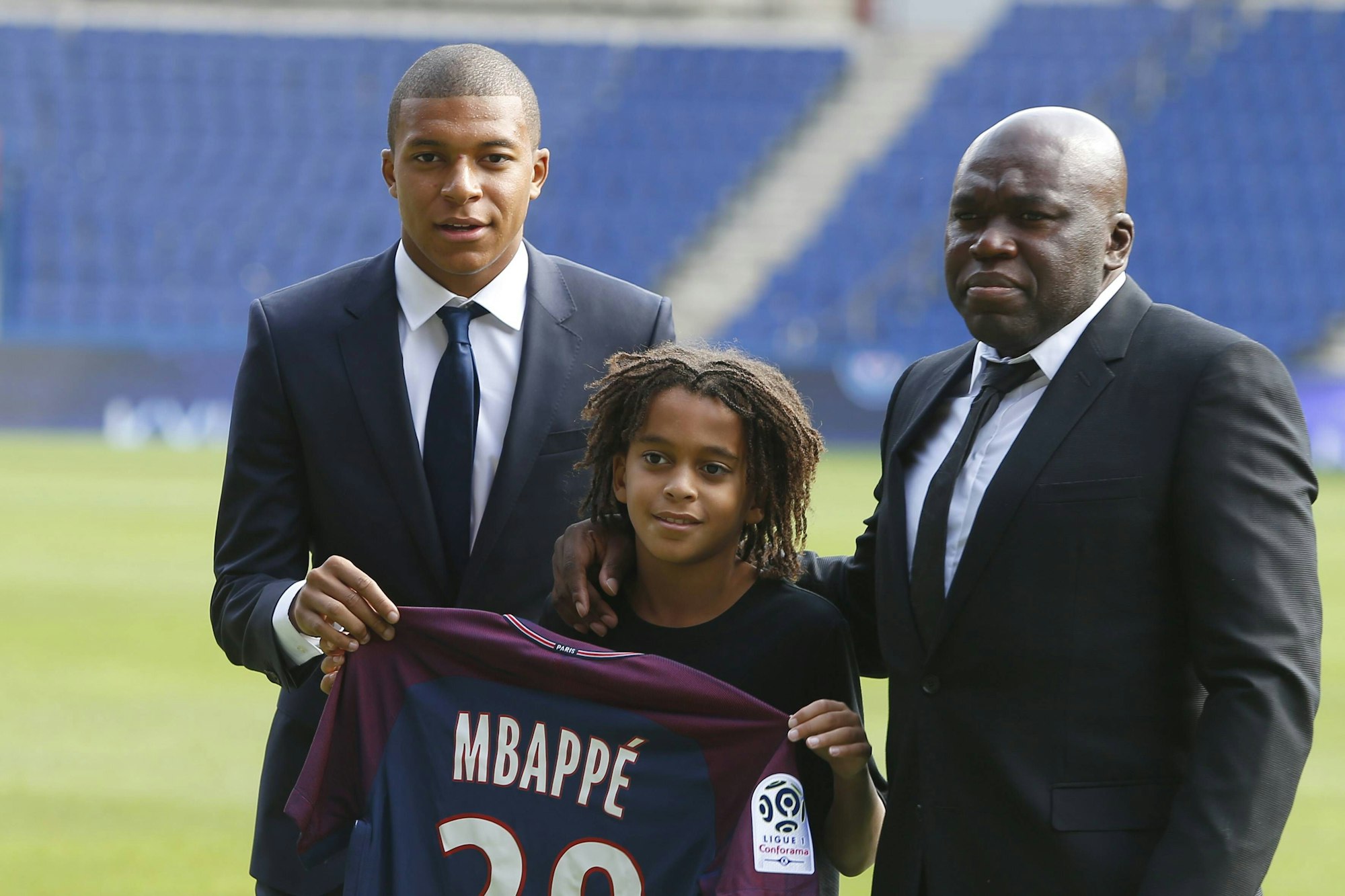 Mbappé hält gemeinsam mit seinem Bruder sein neues Trikot, auch sein Vater gesellt sich dazu und legt eine Hand über die Schulter von Ethan Mbappé.