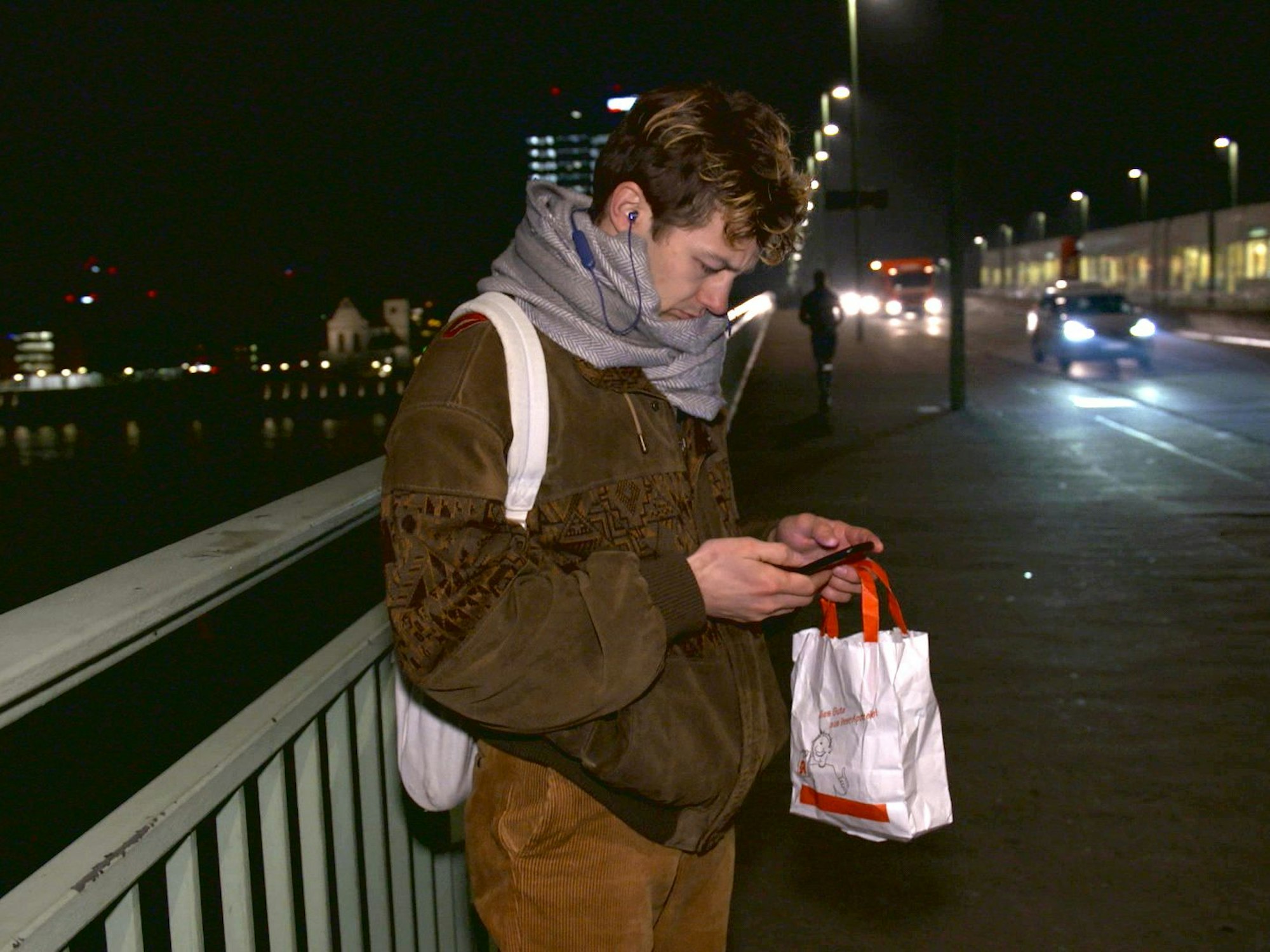 Schauspieler Tim-Fabian Hoffmann steht Nachts auf der Deutzer Brücke und schaut auf sein Handy.
