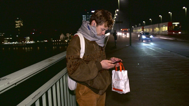 Schauspieler Tim-Fabian Hoffmann steht Nachts auf der Deutzer Brücke und schaut auf sein Handy.