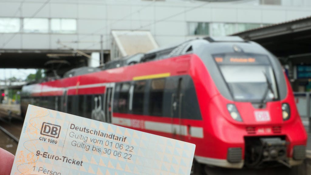 Die Hand eines Mannes hält ein Neun-Euro-Ticket. Auf einem Bahnsteig wartet dabei ein Regionalzug der Deutschen Bahn auf die Abfahrt.