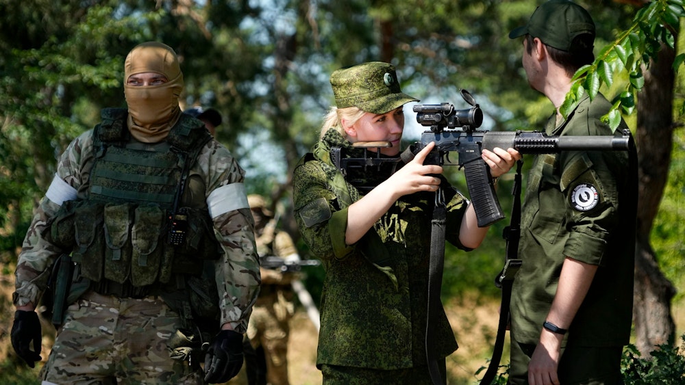 Eine Soldatin der russischen Armee blickt durch ein Scharfschützenzielfernrohr, während sie und andere Soldaten und Soldatinnen eine Gruppe ausländischer Journalisten und Journalistinnen bewachen.
