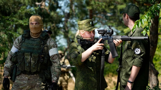 Eine Soldatin der russischen Armee blickt durch ein Scharfschützenzielfernrohr, während sie und andere Soldaten und Soldatinnen eine Gruppe ausländischer Journalisten und Journalistinnen bewachen.