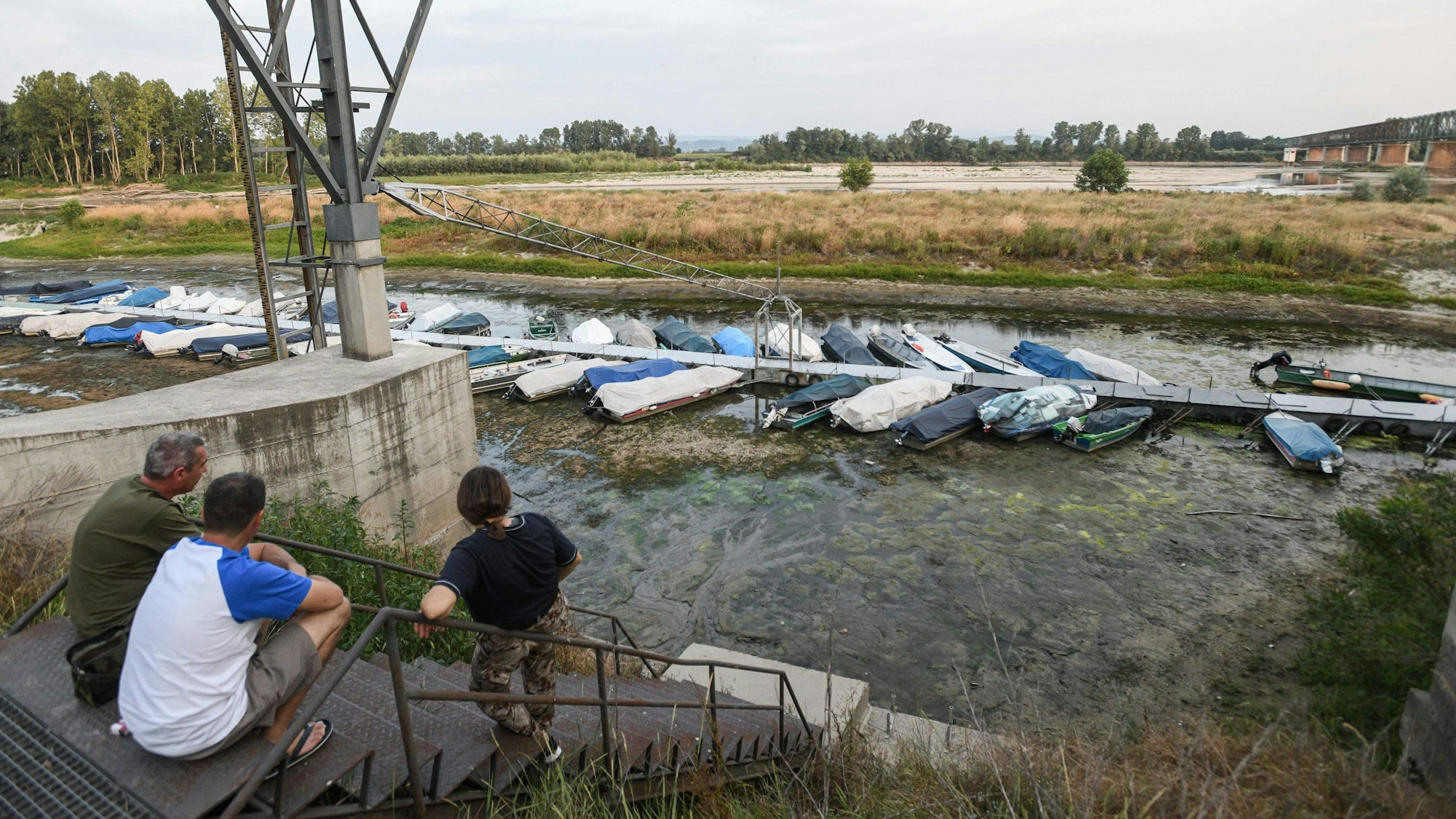 Menschen sitzen bei Ponte della Becca, in der Nähe von Linarolon in der Provinz Pavia in der Lombardei, am 17. Juni 2022 und blicken auf Boote, die bei Niedrigwasser festgemacht sind. In der Region herrscht seit vielen Tagen Dürre und Trockenheit.
