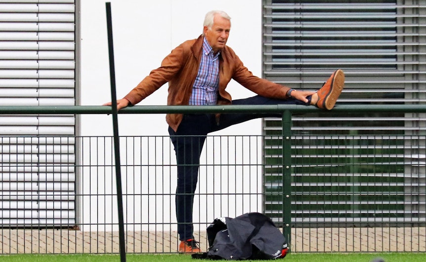 Rainer Bonhof, Vizepräsident von Fußball-Bundeligist Borussia Mönchengladbach, macht während einer Trainingseinheit (27. Juni 2022) der VfL-Profis im Borussia-Park Dehnungsübungen. Bonhof hat sein linkes Bein auf einen Absperrzaun gestreckt.